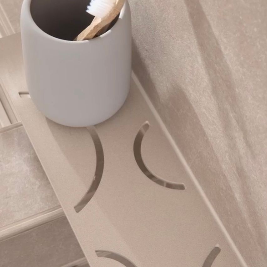 A toothbrush is inside a white cup placed on a bathroom shelf. The shelf has grooves for drainage.