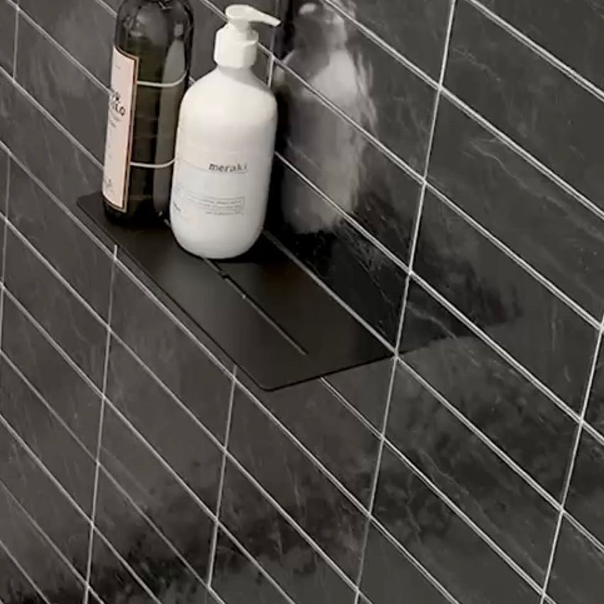 A black shelf with a soap dispenser and a bottle on it is against a black tiled wall.