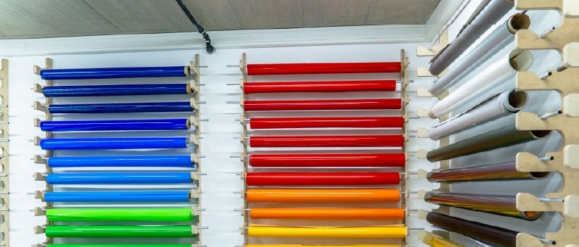 Rollen mit farbigen Plastikfolien sind ordentlich an einer weißen Wand angeordnet, mit blauen, roten, orangefarbenen und grünen Rollen auf der linken Seite und weiteren Rollen auf der rechten Seite.