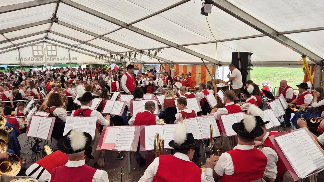 A large group of people in red vests and hats are performing with musical instruments in a tent. The tent has white walls and a black ceiling with lights.