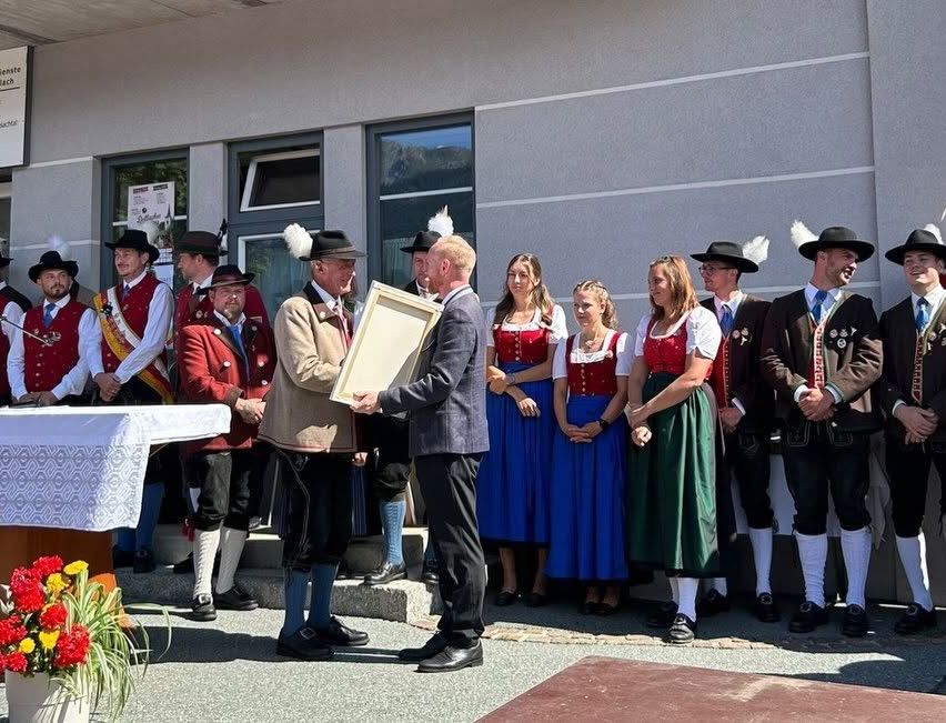 Several people in traditional clothing stand on steps. A man in a suit presents a framed award to a man in a red jacket.