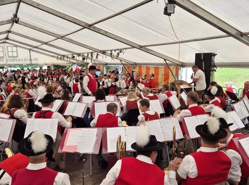 A large orchestra performs on stage under a tent, with musicians dressed in red vests and white shirts, reading music sheets.