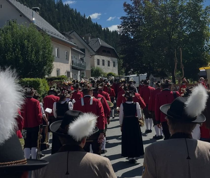 A large group of people in red and white uniforms walk in a parade. They wear hats with white feathers. Some hold musical instruments. Buildings and trees are in the background.