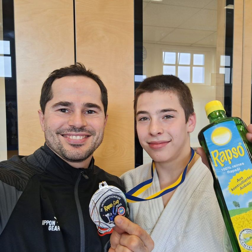 Two men are smiling in a gym, one is wearing a medal and the other is holding a bottle of Rapso oil.