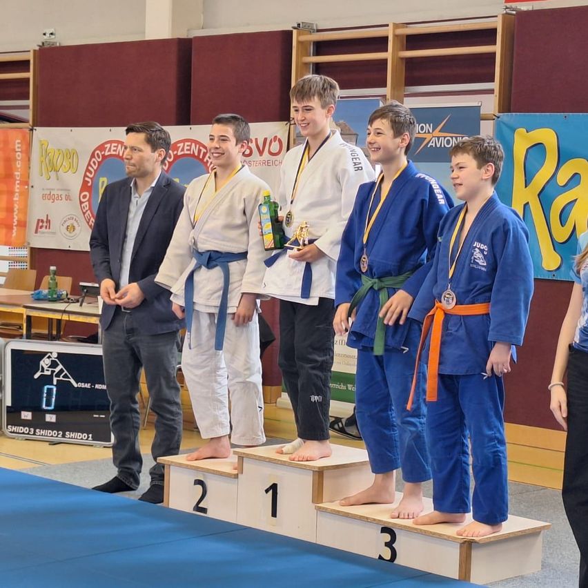 A group of young men in martial arts uniforms stand on a podium with medals around their necks. They are wearing blue and white uniforms. One man holds a trophy.