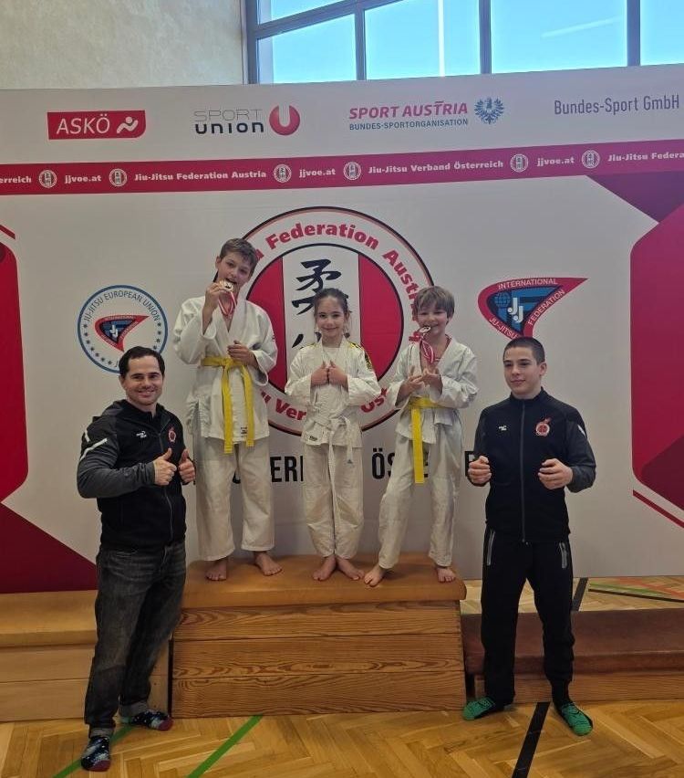 Three young athletes in martial arts uniforms stand on a podium, holding medals. Two are children and one is a man, all wearing black jackets. They are in front of a large banner with logos and text.