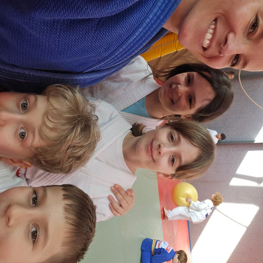 A group of smiling children and a woman, possibly in a martial arts dojo. One child is in a blue uniform and kneeling on the floor.