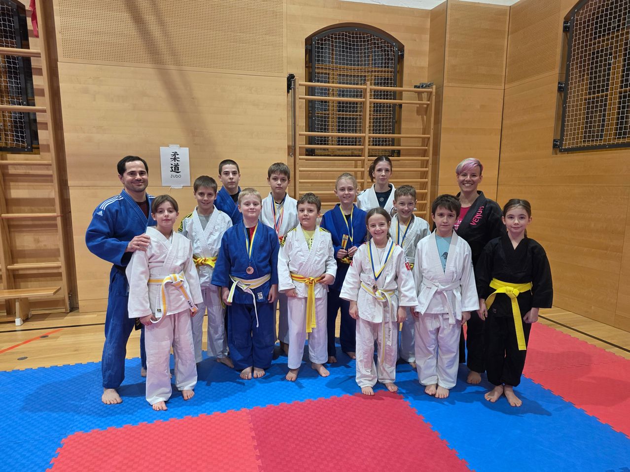 A group of children in martial arts uniforms pose for a photo in a gym, some wearing medals. Behind them, a signboard is mounted on the wall.