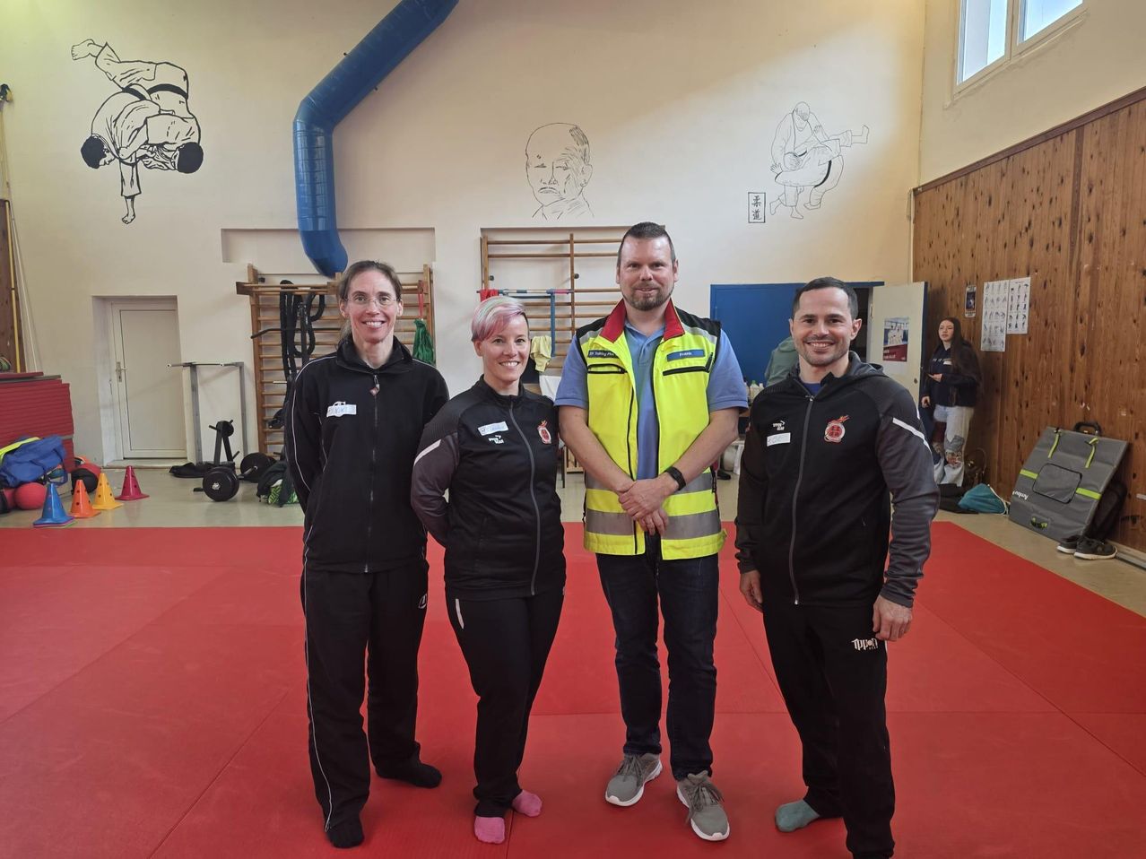 Vier Personen stehen auf einem roten Teppich in einer Turnhalle. Drei Frauen und ein Mann, alle lächelnd und posieren für ein Foto. Der Mann trägt eine Warnweste. Fitnessgeräte und Wandzeichnungen sind im Hintergrund.