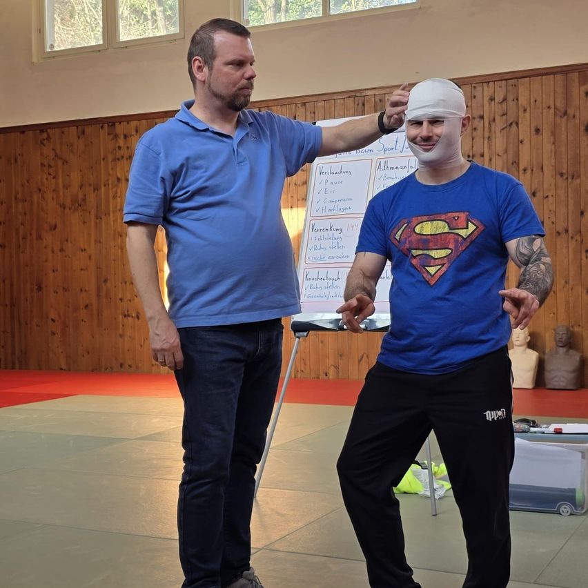 Zwei Männer sind in einem Fitnessstudio. Einer ist in Blau gekleidet und hat einen Verband auf der Stirn. Der andere trägt ein Superman-Shirt. Dahinter steht eine Tafel mit Schrift.