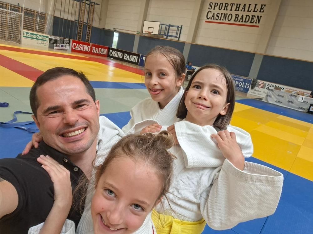 Ein Mann lächelt mit zwei Mädchen in weißen Judouniformen in einer Sporthalle mit gelbem Boden. Sie posieren für ein Foto.