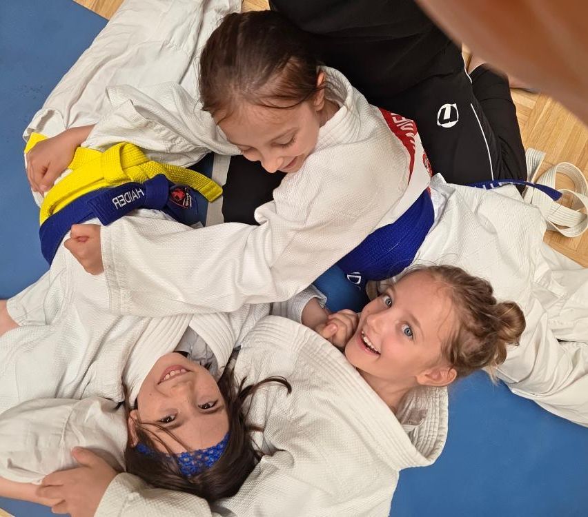 Drei junge Mädchen in weißen Kampfsportuniformen und Gürteln üben auf einem blauen Boden. Das Mädchen in der Mitte hält ein anderes Mädchen auf ihrem Rücken, während das dritte Mädchen ihre Arme hält.
