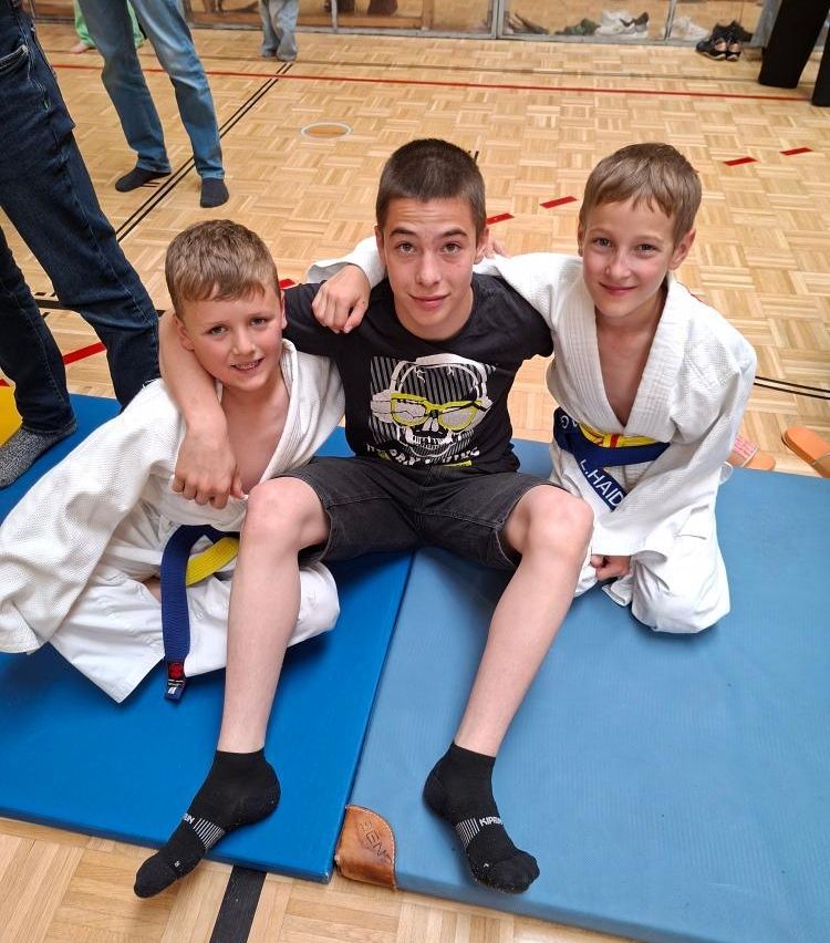 Drei Jungen in Kampfsportuniformen posieren für ein Foto. Einer sitzt auf einer blauen Matte, während die anderen neben ihm sitzen. Sie alle haben lächelnde Gesichter.