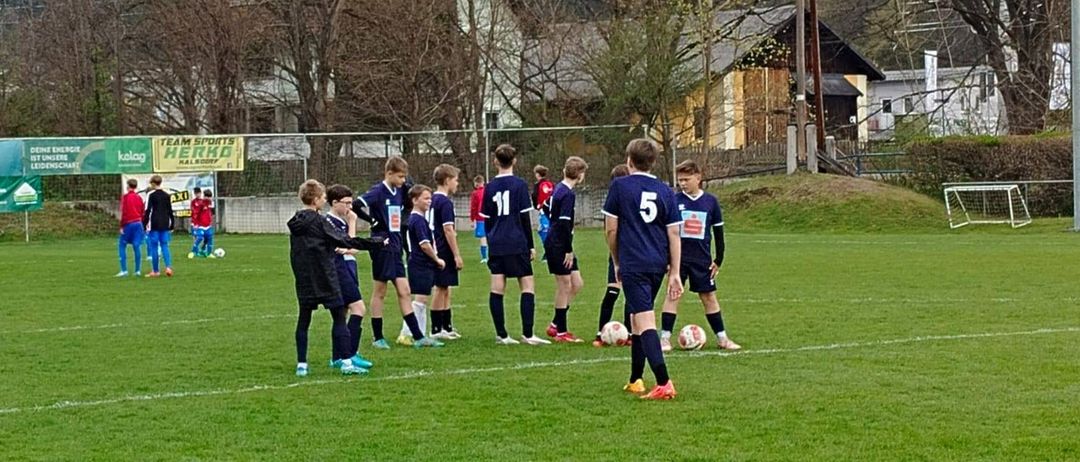 Eine Fußballmannschaft versammelt sich auf einem Feld. Spieler in blauen Trikots stehen mit ihren Fußballbällen. Ein grünes Banner ist im Hintergrund sichtbar.