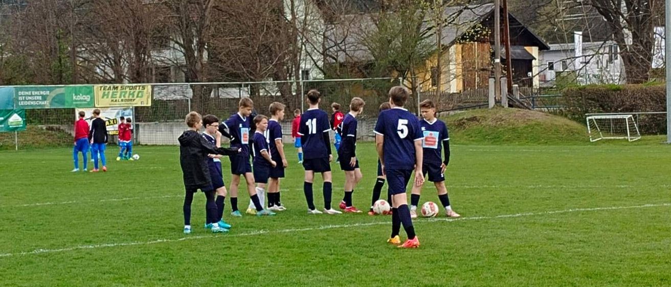 Eine Fußballmannschaft versammelt sich auf einem Feld. Spieler in blauen Trikots stehen mit ihren Fußballbällen. Ein grünes Banner ist im Hintergrund sichtbar.