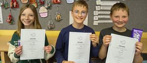 Drei Kinder posieren mit Urkunden für ein Foto. Sie stehen vor einer Pinnwand mit Cupcakes und anderen Dekorationen. Der Junge in der Mitte trägt eine Brille.