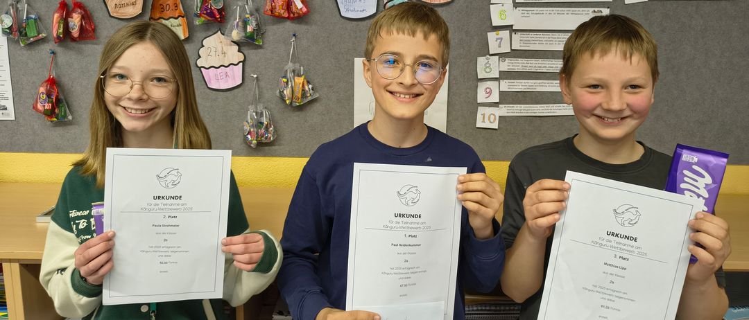 Drei Kinder posieren mit Urkunden für ein Foto. Sie stehen vor einer Pinnwand mit Cupcakes und anderen Dekorationen. Der Junge in der Mitte trägt eine Brille.