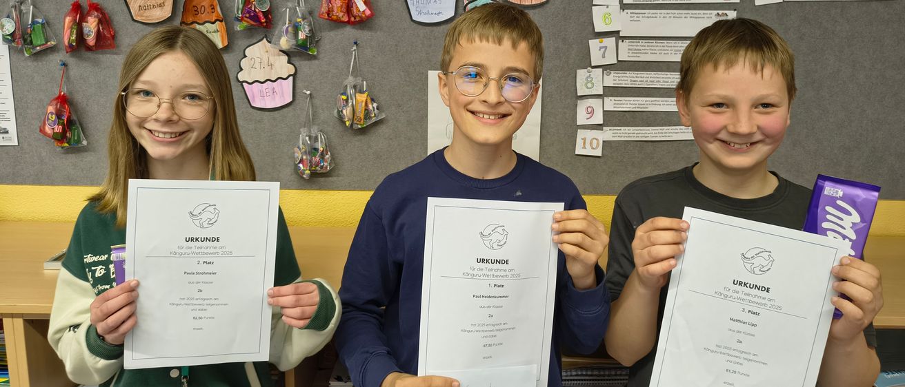 Drei Kinder posieren mit Urkunden für ein Foto. Sie stehen vor einer Pinnwand mit Cupcakes und anderen Dekorationen. Der Junge in der Mitte trägt eine Brille.