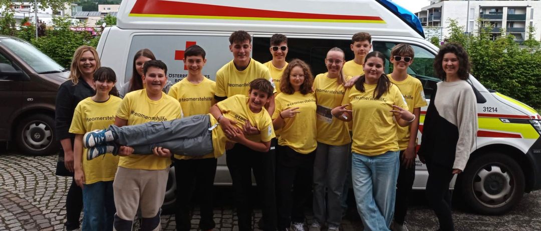 Eine Gruppe junger Menschen in gelben Shirts mit dem Text '#teamerstehilfe' steht vor einem Krankenwagen. Sie posieren lächelnd für ein Foto.