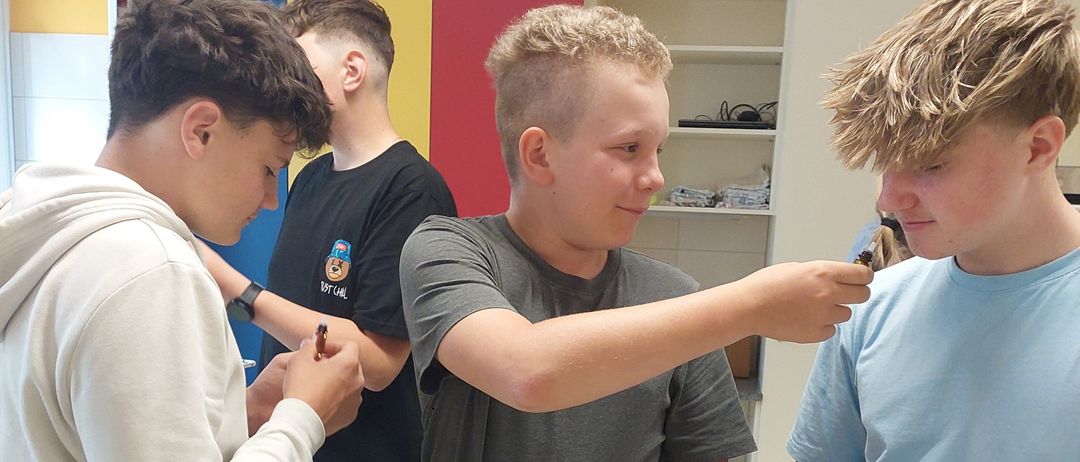 Vier Jungen sind in einem Labor und handhaben Gummibärchen. Ein Junge zeigt auf einen Gummibär und ein anderer hält eine Flasche. Der Tisch ist mit Flaschen, einem gelben Schneidebrett und einem Papier übersät.