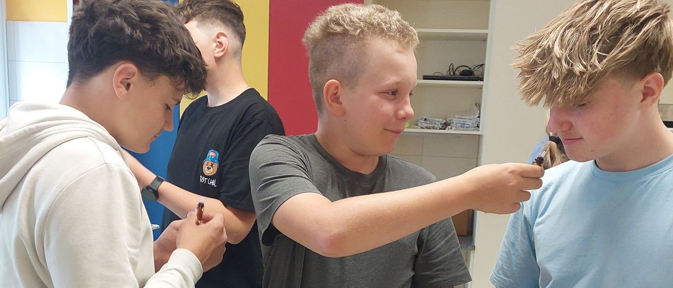 Vier Jungen sind in einem Labor und handhaben Gummibärchen. Ein Junge zeigt auf einen Gummibär und ein anderer hält eine Flasche. Der Tisch ist mit Flaschen, einem gelben Schneidebrett und einem Papier übersät.