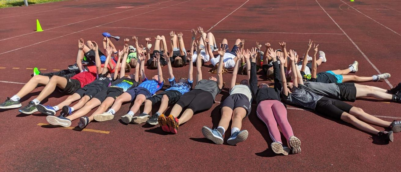 Eine Gruppe von Menschen liegt auf einer Laufbahn, hebt die Hände und trägt Turnschuhe. Einige tragen Armbänder. Auf der Laufbahn sind Kegel platziert.