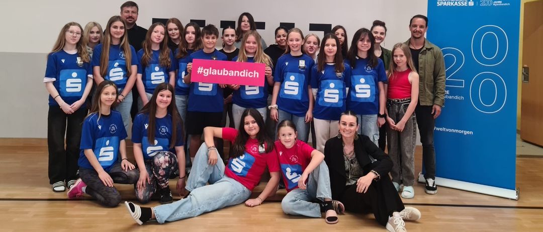 Eine Gruppe junger Menschen in blauen Trikots posiert für ein Foto in einer Turnhalle mit einem Basketballkorb und einem Banner mit der Aufschrift '20'.