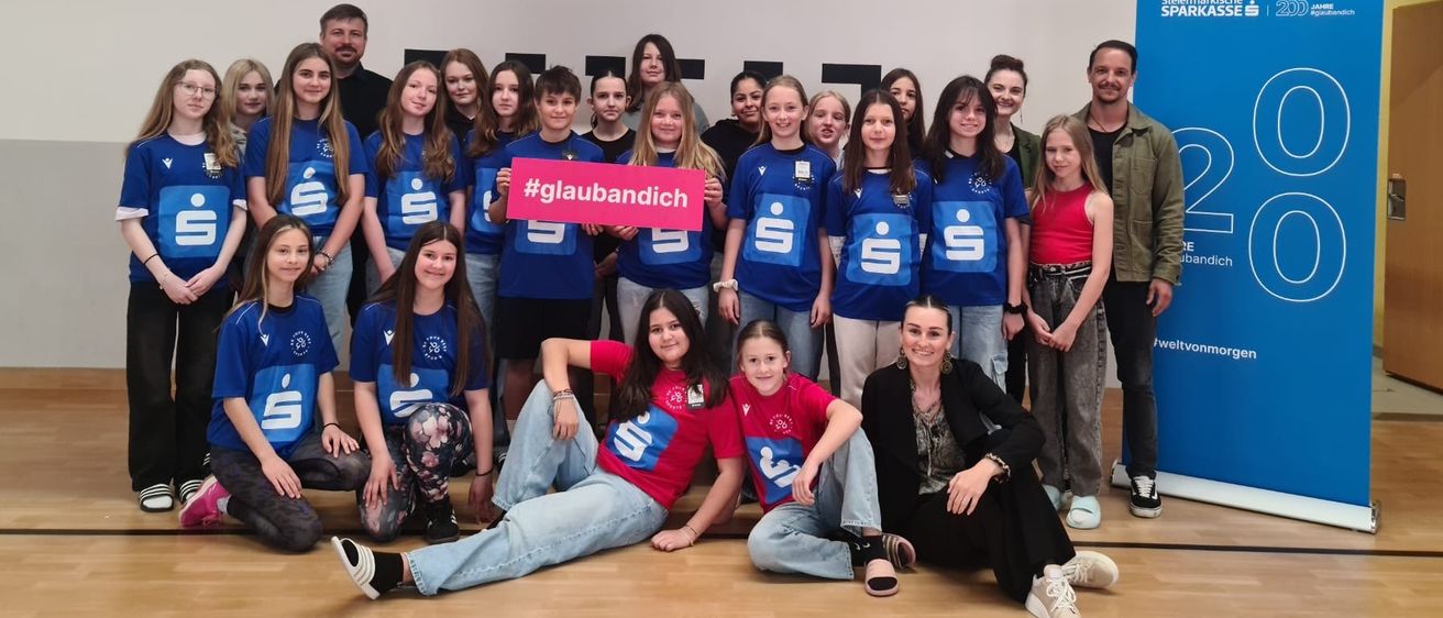 Eine Gruppe junger Menschen in blauen Trikots posiert für ein Foto in einer Turnhalle mit einem Basketballkorb und einem Banner mit der Aufschrift '20'.