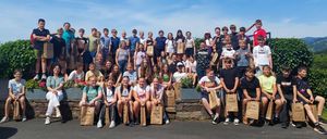 Eine große Gruppe von Menschen, möglicherweise Schüler, versammelt sich draußen und hält Papiertüten in der Hand. Sie stehen und sitzen auf einer Betonmauer mit einem Busch dahinter. Einige posieren für ein Foto an einem sonnigen Tag.