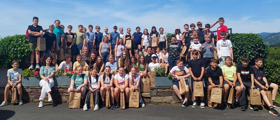 Eine große Gruppe von Menschen, möglicherweise Schüler, versammelt sich draußen und hält Papiertüten in der Hand. Sie stehen und sitzen auf einer Betonmauer mit einem Busch dahinter. Einige posieren für ein Foto an einem sonnigen Tag.
