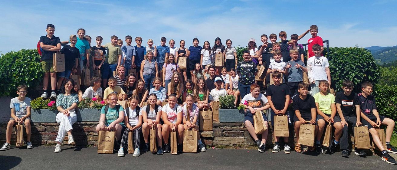 Eine große Gruppe von Menschen, möglicherweise Schüler, versammelt sich draußen und hält Papiertüten in der Hand. Sie stehen und sitzen auf einer Betonmauer mit einem Busch dahinter. Einige posieren für ein Foto an einem sonnigen Tag.