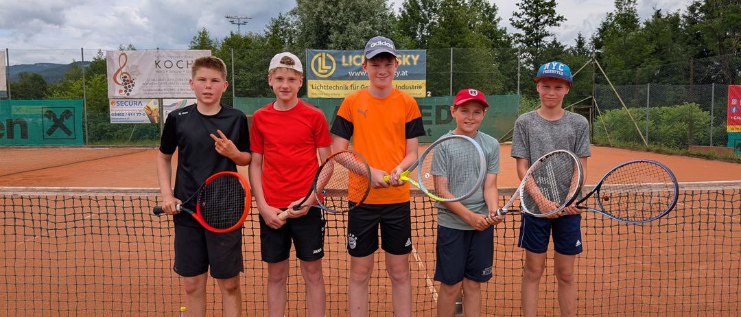 Fünf Jungen stehen auf einem Tennisplatz mit Tennisschlägern. Sie tragen verschiedenfarbige Hemden und Shorts. Hinter ihnen steht ein Zaun mit Bannern. Bäume und ein bewölkter Himmel sind im Hintergrund.