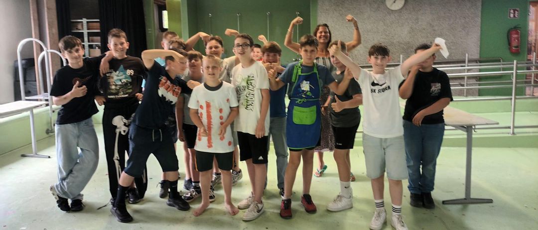 Eine Gruppe junger Jungen und eine Frau posieren für ein Foto in einem Raum mit grünen Wänden. Einige von ihnen tragen Schürzen und Turnschuhe.