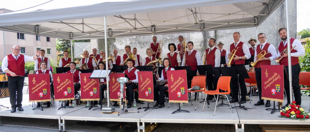 Eine Gruppe von Musikern, gekleidet in rote Westen, tritt auf einer Bühne unter einem weißen Zelt auf, mit roten Bannern hinter ihnen.