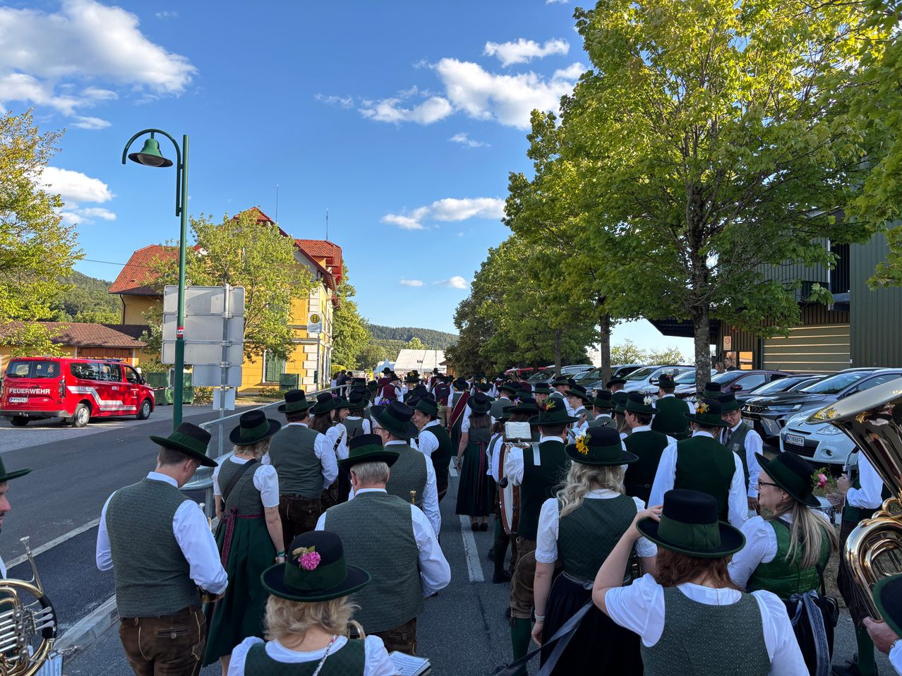 Eine große Gruppe von Menschen in traditioneller bayerischer Kleidung, einschließlich grüner Westen und Hüte, geht eine von Bäumen und geparkten Autos gesäumte Straße entlang. Der Himmel ist blau mit einigen Wolken.