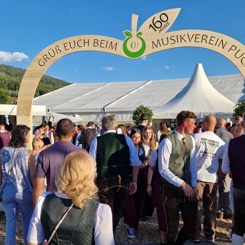 Eine Menschenmenge in traditioneller Kleidung versammelt sich unter einem Holzbogen mit der Aufschrift '160 Jahre Musikverein Puch'. Der Bogen befindet sich vor einem weißen Zelt.