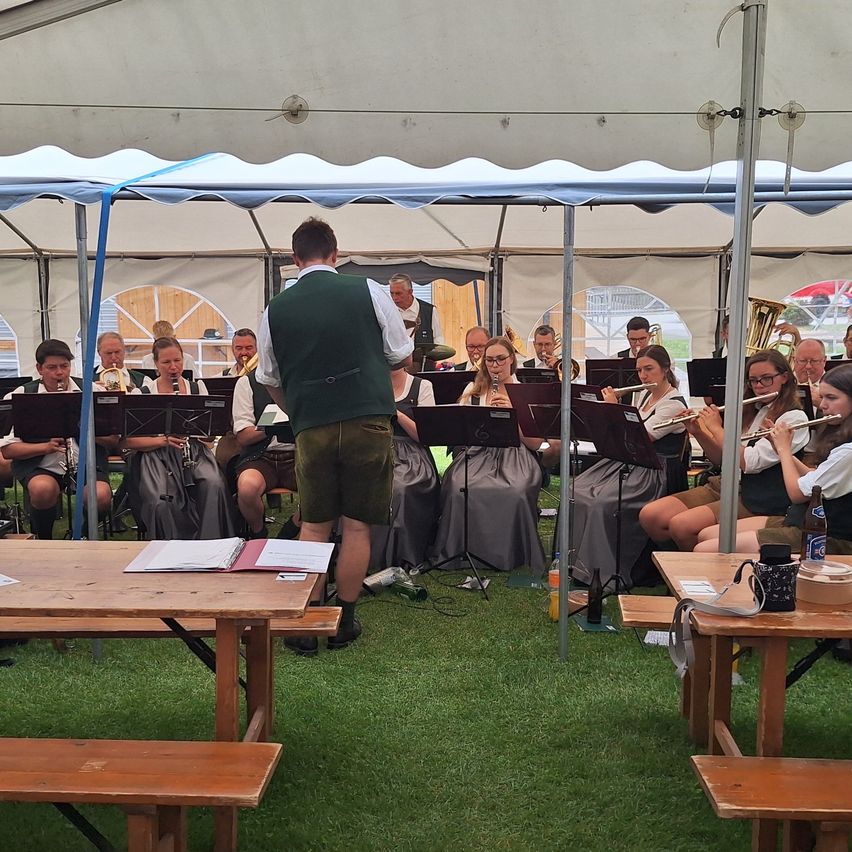 Ein Orchester spielt auf einem Rasenfeld unter einem weißen Zelt. Die Bandmitglieder spielen verschiedene Instrumente, und ein Mann steht vor ihnen.