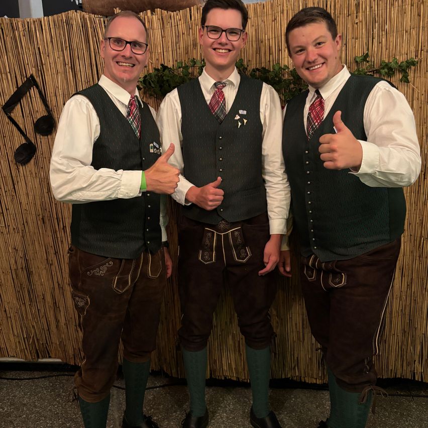 Three men dressed in traditional German attire, standing together and giving thumbs up in front of a bamboo wall.