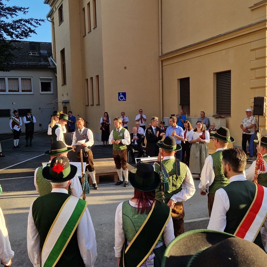A group of people dressed in traditional attire stand in front of a building, with others watching from behind. The setting appears to be a cultural event.