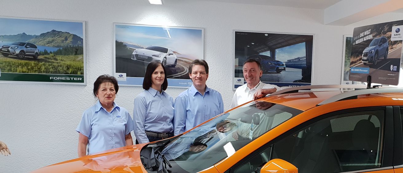 Drei Personen stehen neben einem orangefarbenen Auto in einer Ausstellung. Das Auto hat ein Dachträgersystem und ein Schild. Sie tragen passende hellblaue Hemden. Hinter ihnen hängen Plakate von Autos.