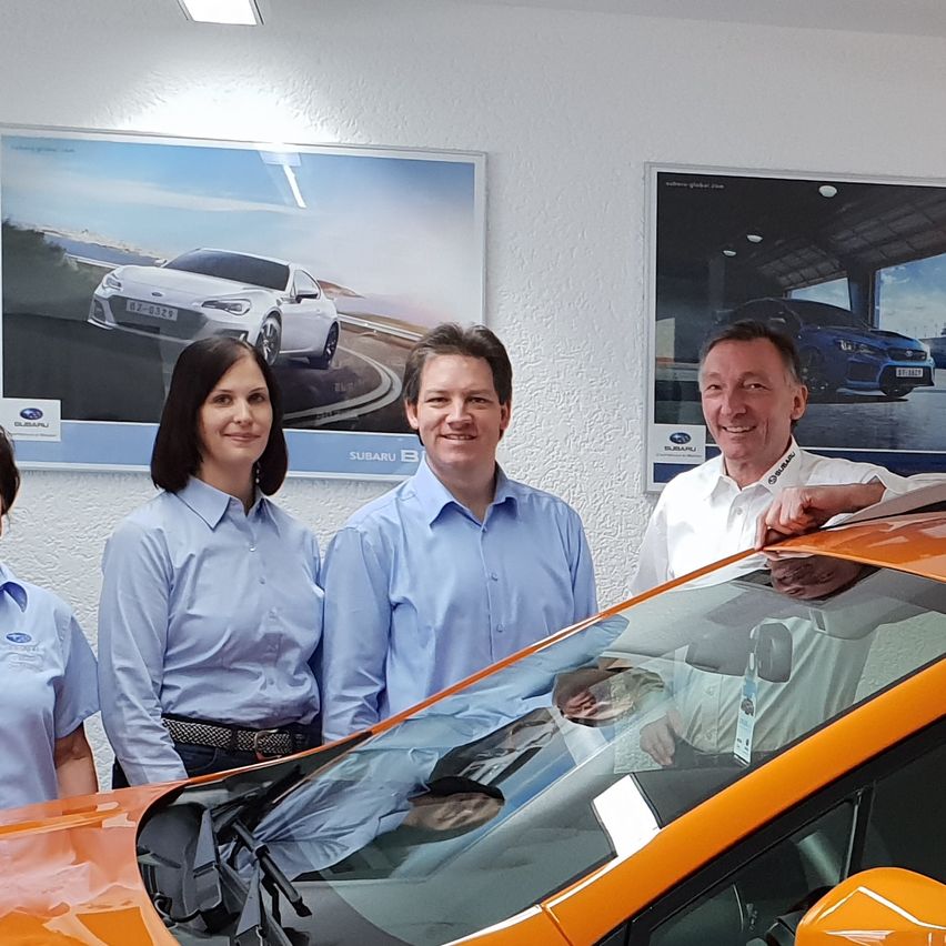 Drei Personen stehen neben einem orangefarbenen Auto in einer Ausstellung. Das Auto hat ein Dachträgersystem und ein Schild. Sie tragen passende hellblaue Hemden. Hinter ihnen hängen Plakate von Autos.