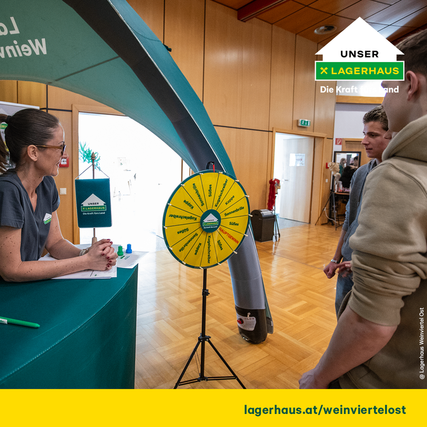 Eine Frau mit Brille sitzt an einem Schreibtisch vor einem Drehrad, zwei Männer stehen in der Nähe, die Umgebung ist innen mit Holzböden und einem weißen Pfeilschild.