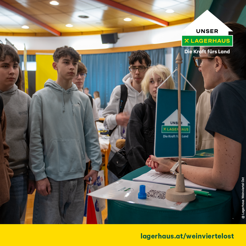 Eine Gruppe junger Menschen steht um einen Tisch mit einem Schild, auf dem 'Unser Lagerhaus' steht. Eine Frau spricht mit ihnen. Hinter ihr befindet sich ein großes Banner und Menschen mit Brille. Der Boden ist aus Holz.