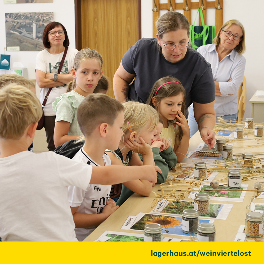 A group of children are gathered around a table with jars of seeds and plants, guided by a woman in a classroom.