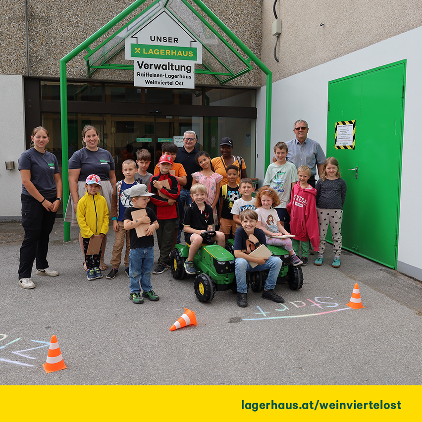 A group of children and adults pose for a photo in front of a building with a sign that reads Unser Lagerhaus Verwaltung. The children are sitting on toy tractors and holding papers.