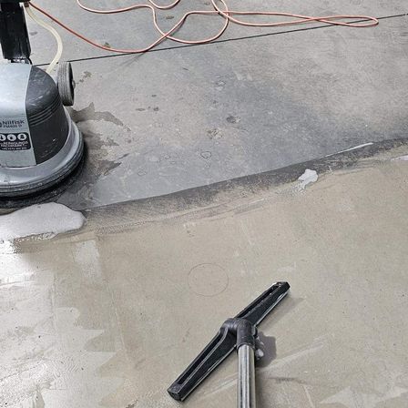 Eine Bodenreinigungsmaschine mit einem Schlauch steht auf einem Betonboden, mit einem Besen in der Nähe und einem nassen Bereich.