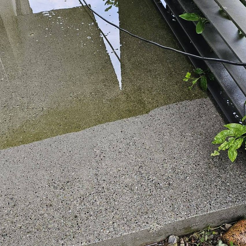 Eine Nahaufnahme einer Betonoberfläche mit der Reflexion eines Gebäudes und einem schwarzen Kabel. Der Beton ist nass und hat einige kleine Löcher.