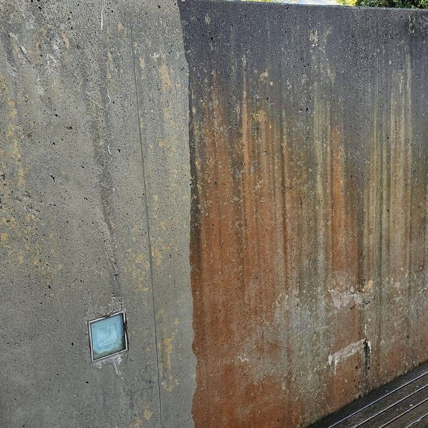 Eine Betonwand mit einem verrosteten Abschnitt und einer kleinen quadratischen Lichtvorrichtung auf der linken Seite.