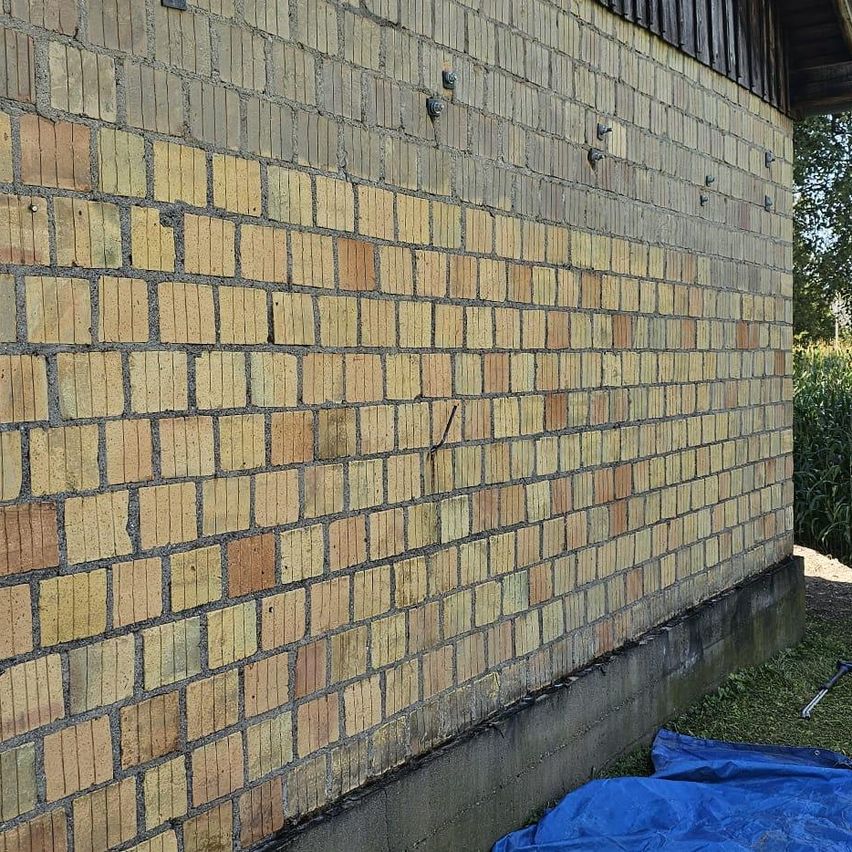 Eine Ziegelwand mit einer Mischung aus gelben und braunen Ziegeln, die Anzeichen von Abnutzung zeigt, wobei einige Mörtelfugen sichtbar sind. Ein blauer Tarp liegt vor der Wand auf dem Boden.