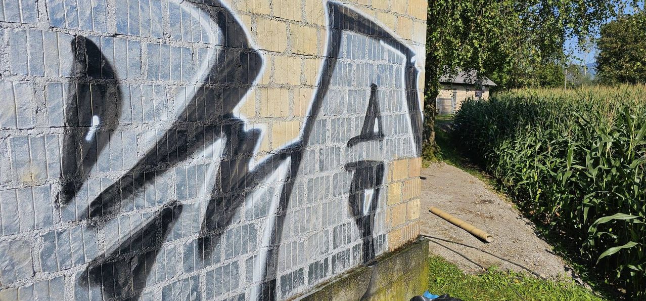 Eine Ziegelmauer mit schwarzen Graffiti, einschließlich eines großen Buchstabens 'A' und einiger anderer Buchstaben, in einer Außenumgebung.
